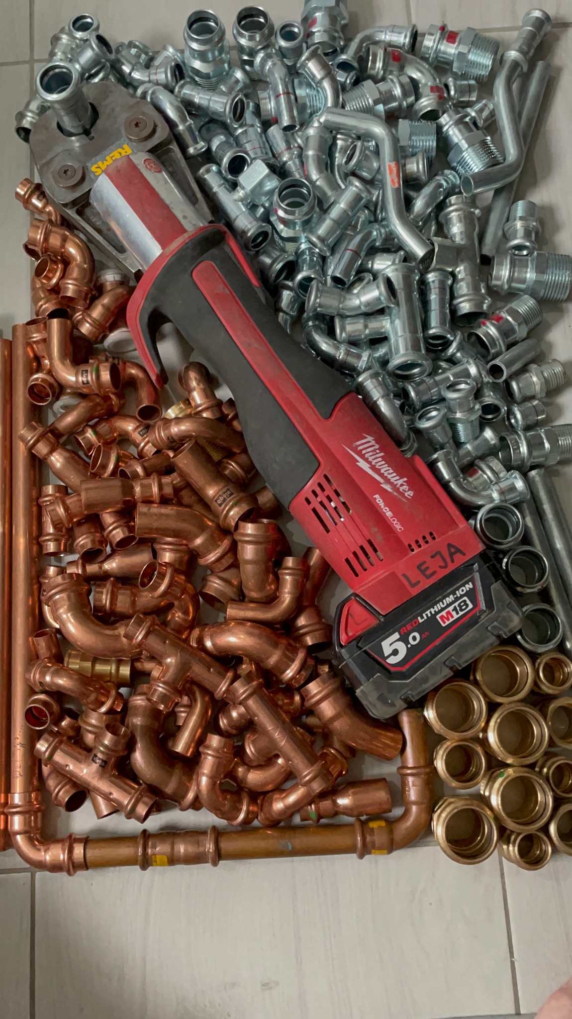 Stos miedzianych i srebrnych kształtek hydraulicznych oraz zaciskarki akumulatorowej Milwaukee z baterią 5.0 Ah na jasnym tle.