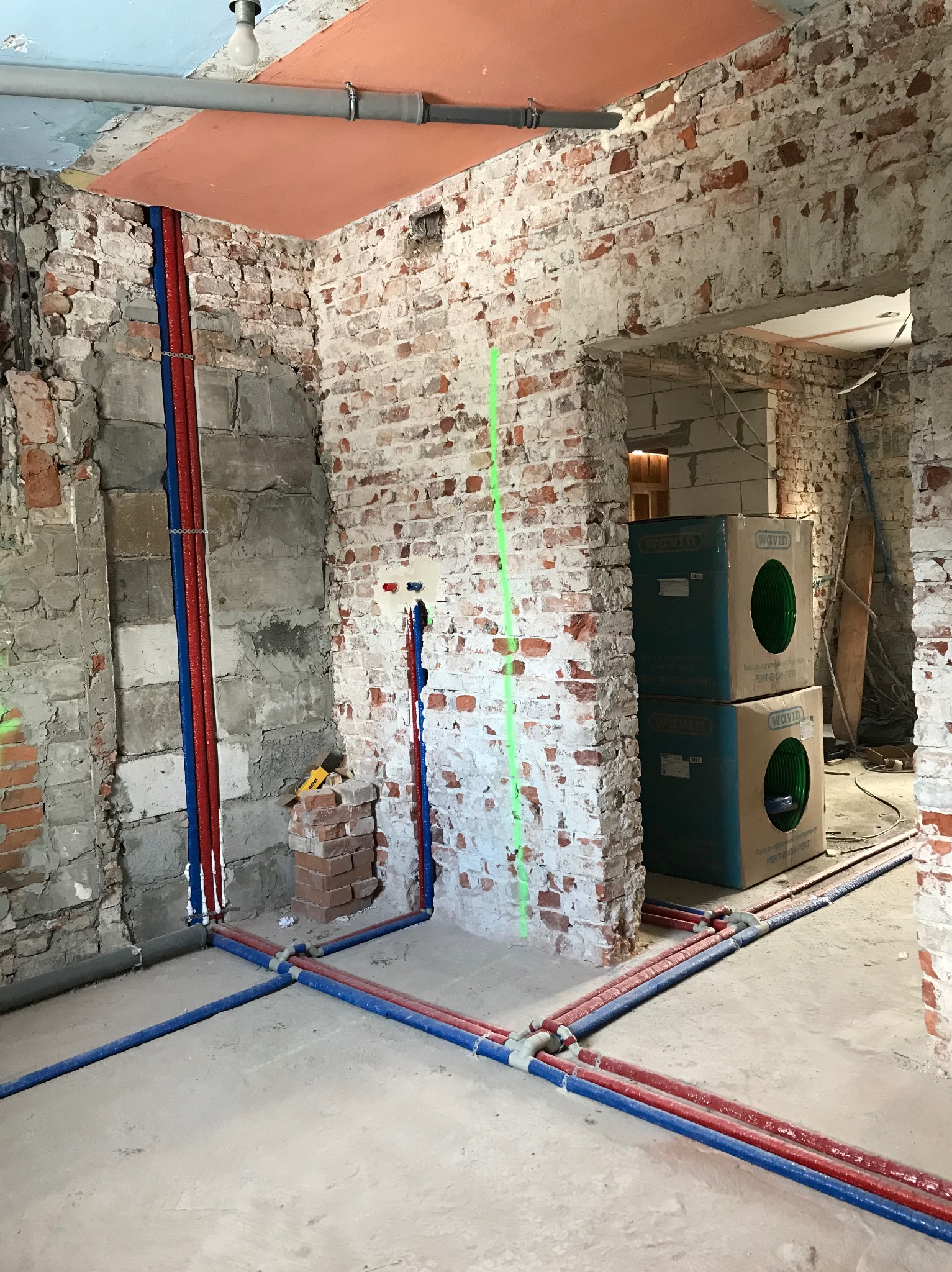 Wnętrze w trakcie remontu z widocznymi nowymi instalacjami hydraulicznymi: rurami czerwonymi i niebieskimi, częściowo ukrytymi w ścianie z surowej cegły, oraz dwoma kartonami Wavin w tle.