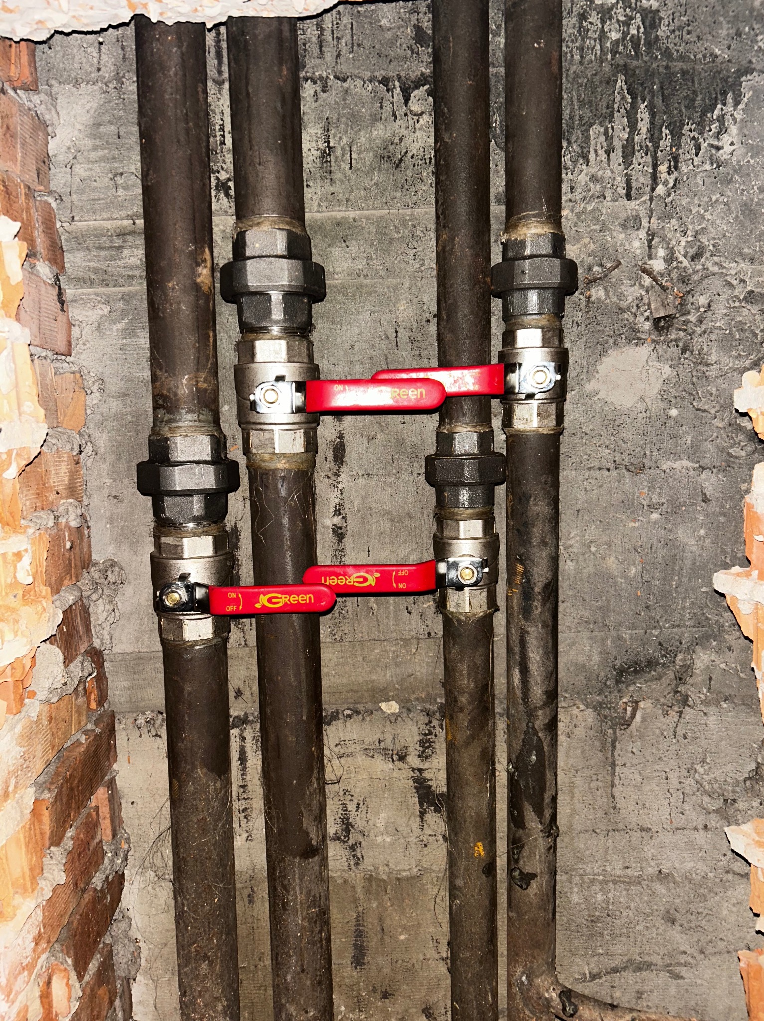 Dwie pary starych, pionowych rur z zaworami odcinającymi JGreen z czerwonymi rączkami, zamontowane przy surowej ścianie z cegły i betonu.
