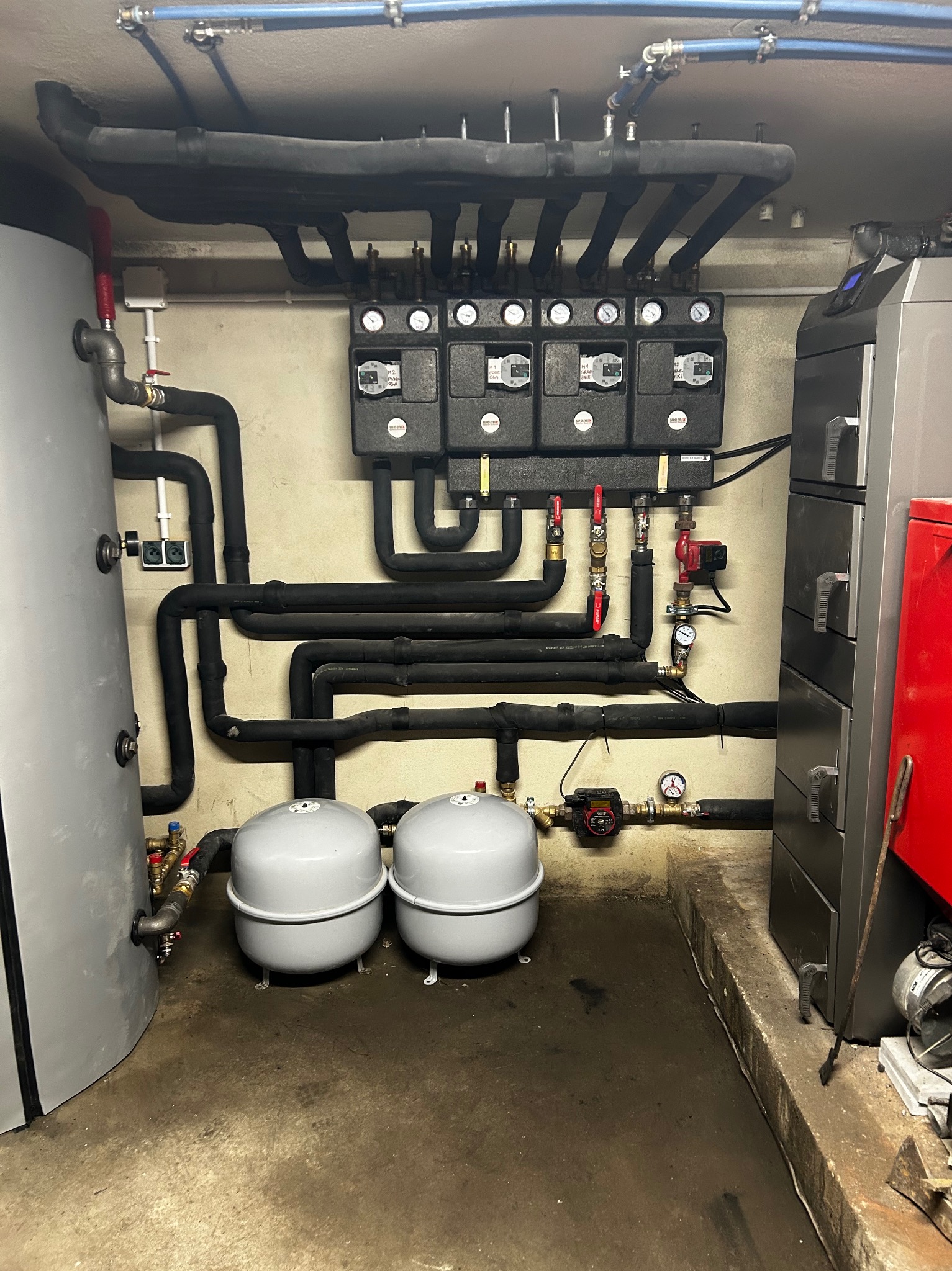 Rozbudowana instalacja grzewcza w kotłowni z czarnymi rurami izolowanymi, dwoma szarymi zbiornikami wyrównawczymi i rozdzielaczami z manometrami, widoczny piec z czerwonym elementem.