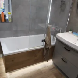 Nowoczesna łazienka z wanną obudowaną drewnem, szklanym parawanem i szarą szafką pod umywalką z roślinnym motywem na boku.