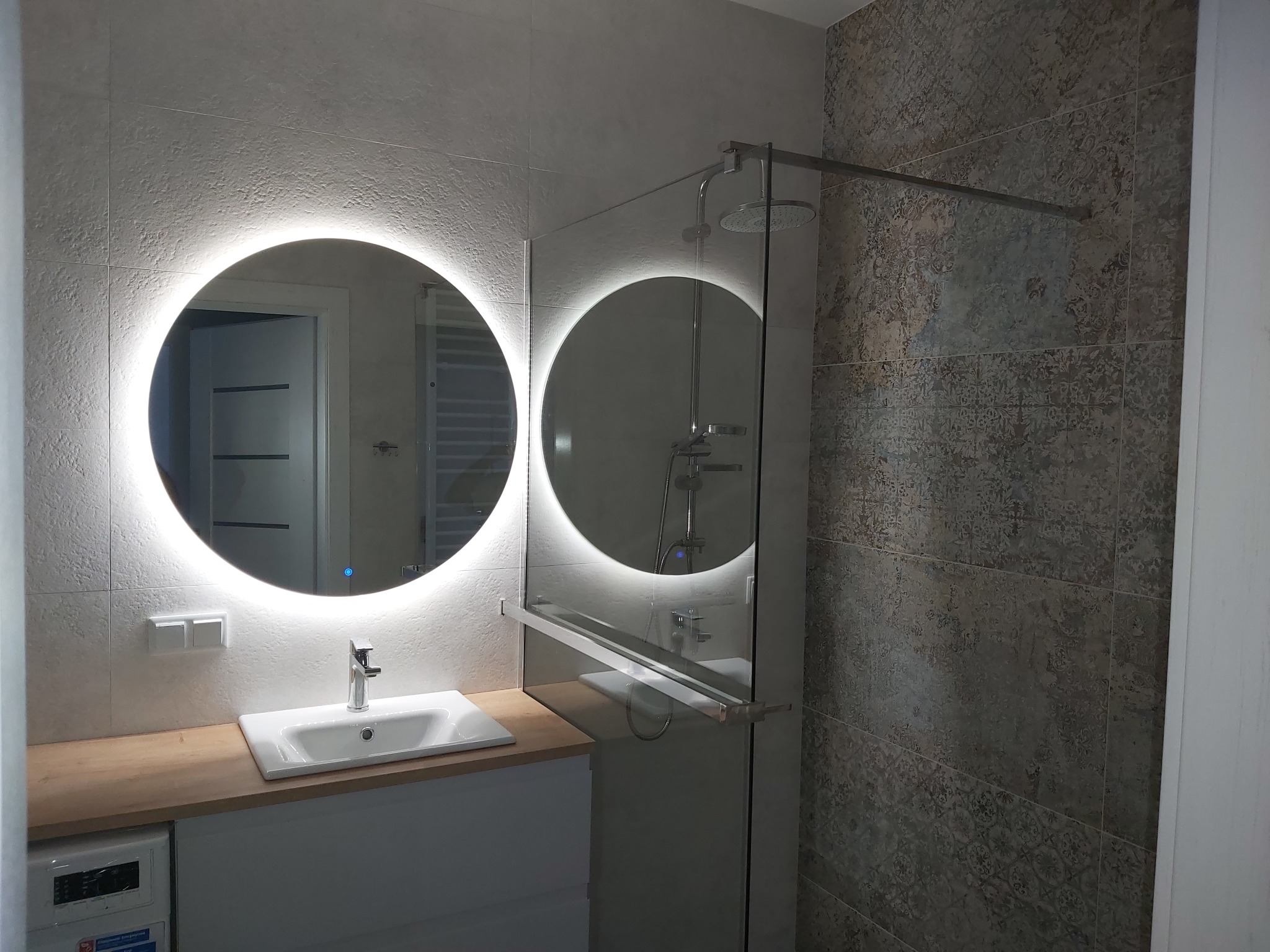 Nowoczesna łazienka z okrągłym lustrem podświetlanym LED, białą umywalką nablatową, drewnianym blatem, kabiną prysznicową typu walk-in i płytkami w dwóch odcieniach szarości.