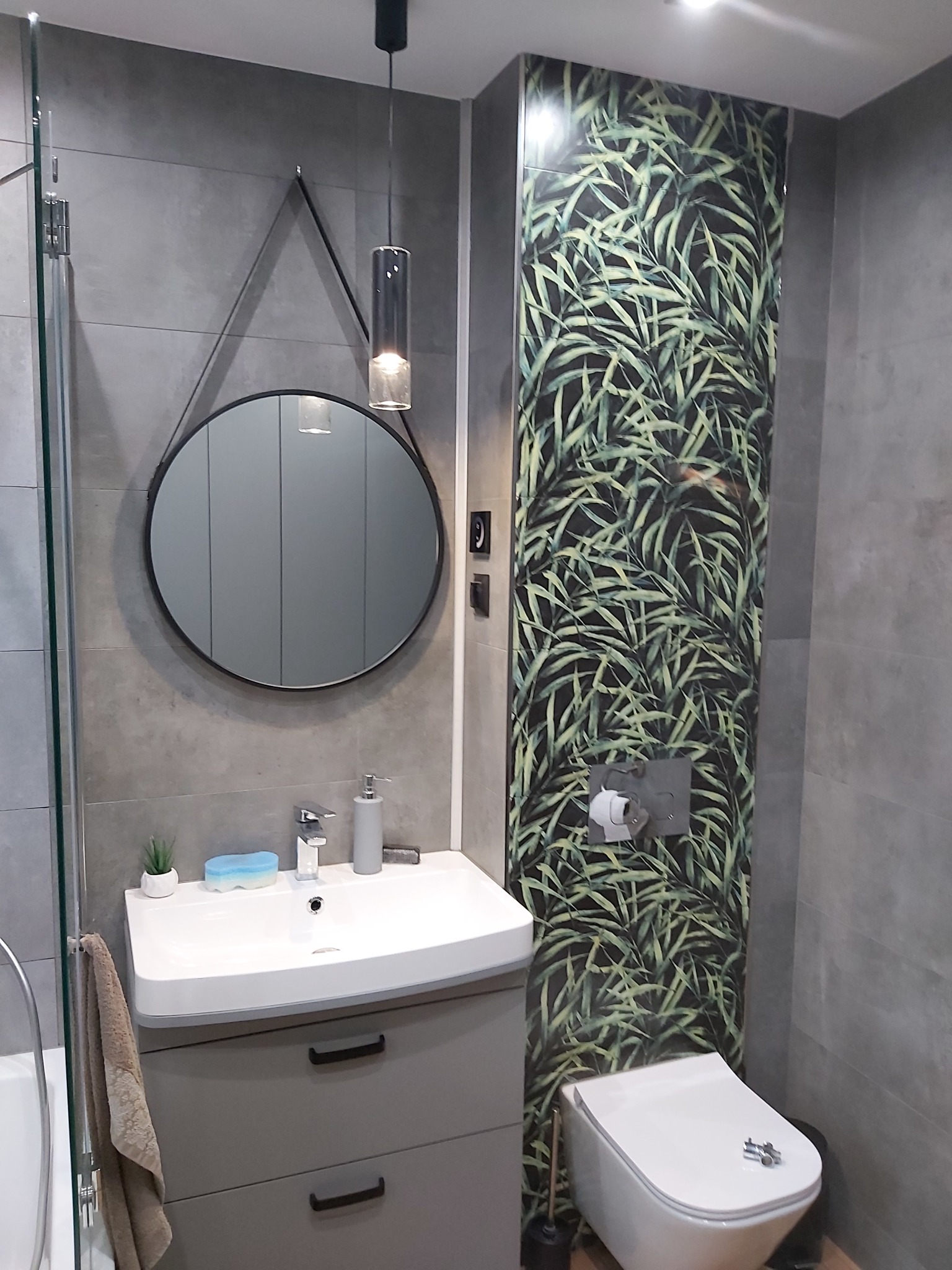 Nowoczesna łazienka z szarymi płytkami, okrągłym lustrem zawieszonym na pasku, umywalką z szafką w kolorze taupe i toaletą obok pionowego panelu z motywem liści palmowych.