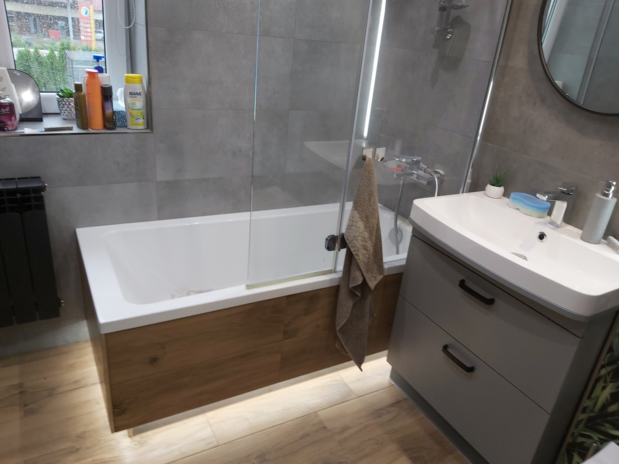 Nowoczesna łazienka z wanną obudowaną drewnem, szklanym parawanem i szarą szafką pod umywalką z roślinnym motywem na boku.