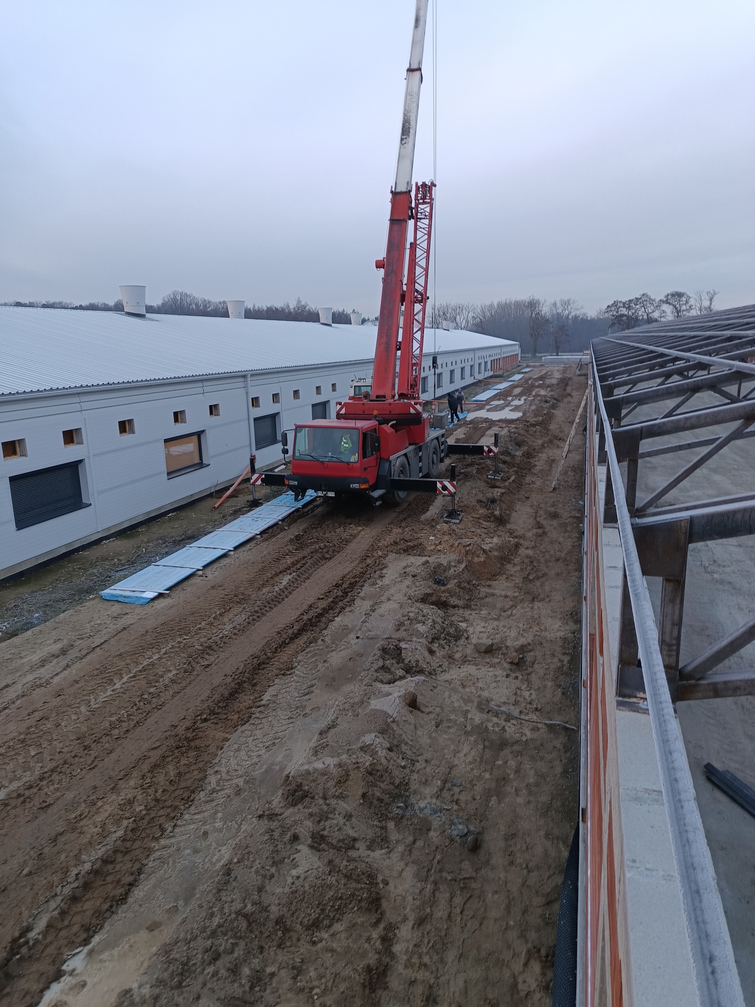 Montaż konstrukcji dachu hali przemysłowej z użyciem dźwigu samochodowego. Widoczne elementy konstrukcyjne i budynek w tle. Pochmurny dzień.