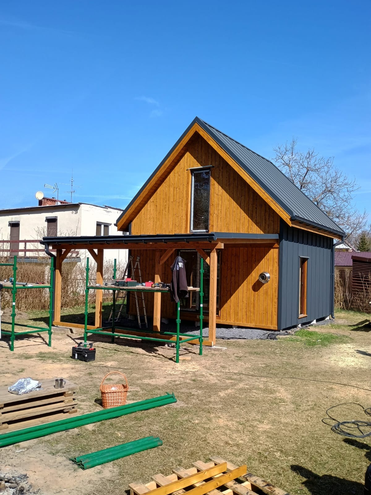 Nowoczesny dom szkieletowy z drewnianą elewacją i szarą częścią, widoczny z zewnątrz, z rusztowaniami i narzędziami sugerującymi trwające prace wykończeniowe.