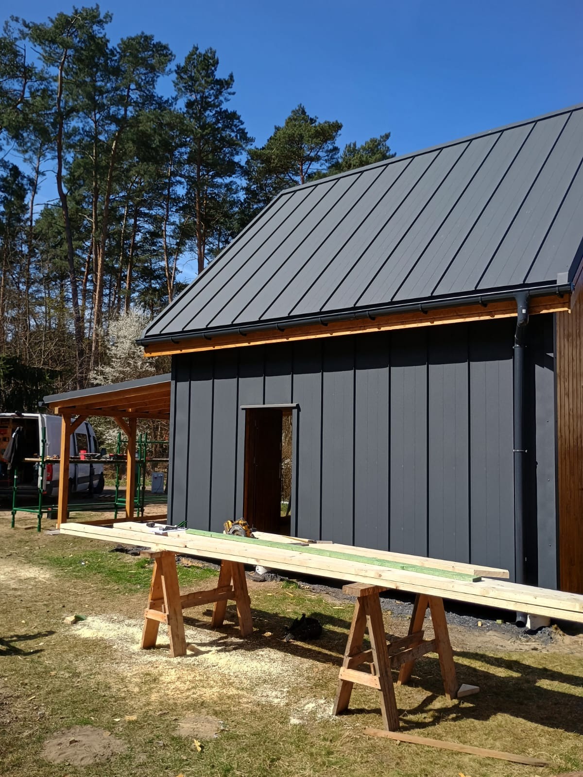 Nowoczesny dom szkieletowy w trakcie budowy: szara elewacja z metalowym dachem, drewniana konstrukcja zadaszenia, deski na kozłach, słoneczny dzień.