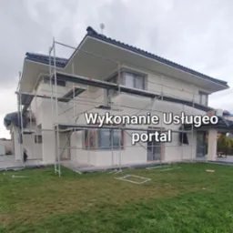 Nowoczesny dom jednorodzinny w trakcie prac wykończeniowych elewacji, otoczony rusztowaniami i zabezpieczony folią malarską.