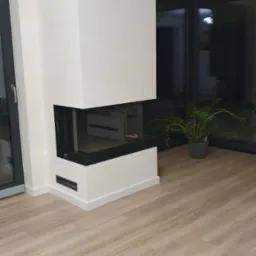 Nowoczesny, narożny kominek z szybą z trzech stron, obudowany białymi płytami z czarnymi wstawkami wentylacyjnymi, na podłodze panele w odcieniach beżu, w tle duże okno i roślina doniczkowa.