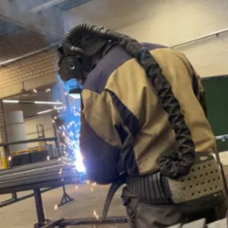 Pracownik w kombinezonie spawalniczym i masce ochronnej podczas spawania metalowych element&oacute;w konstrukcyjnych na warsztacie.