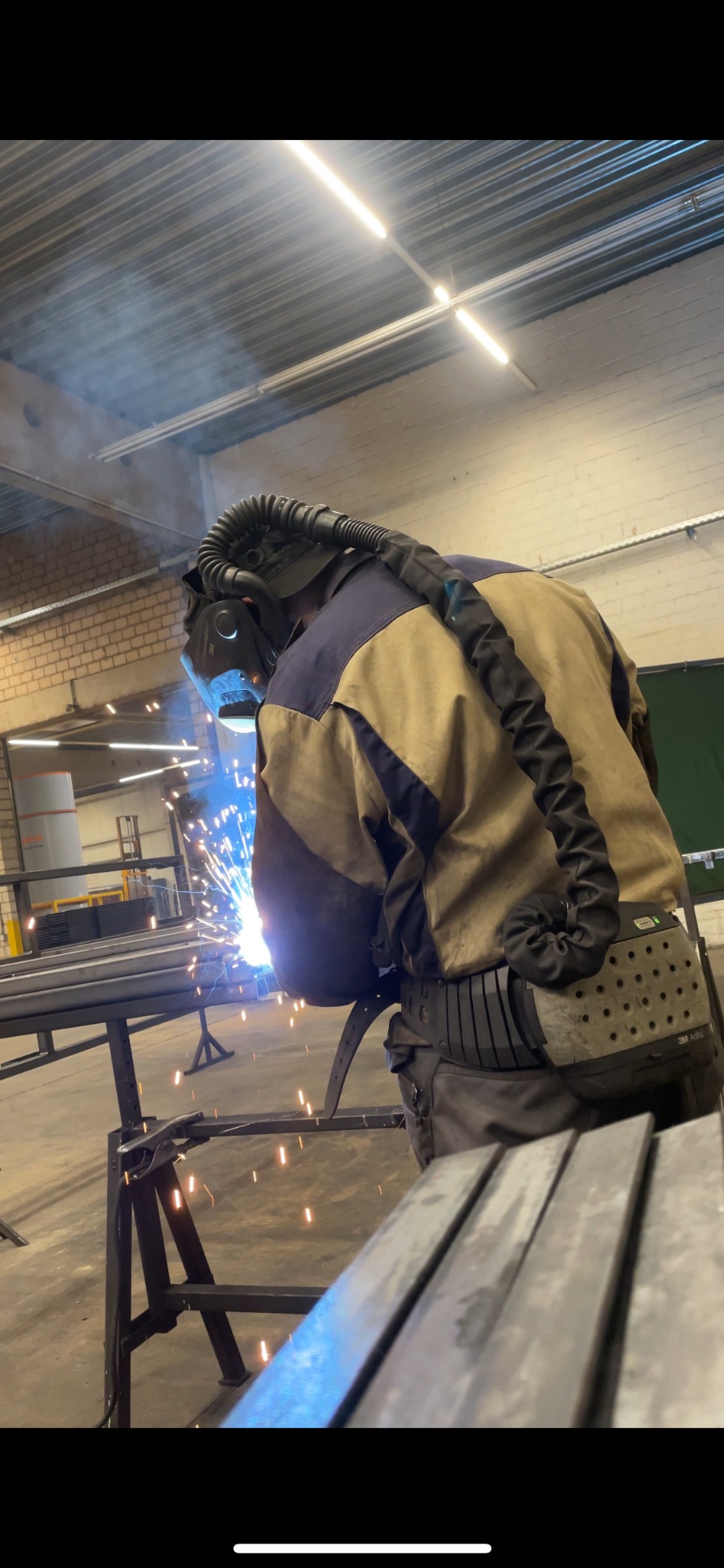 Pracownik w kombinezonie spawalniczym i masce ochronnej podczas spawania metalowych elementów konstrukcyjnych na warsztacie.