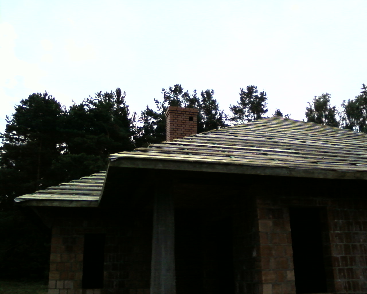 Budowa domu w stanie surowym otwartym z widocznym kominem z cegły i przygotowanym dachem, otoczony drzewami.