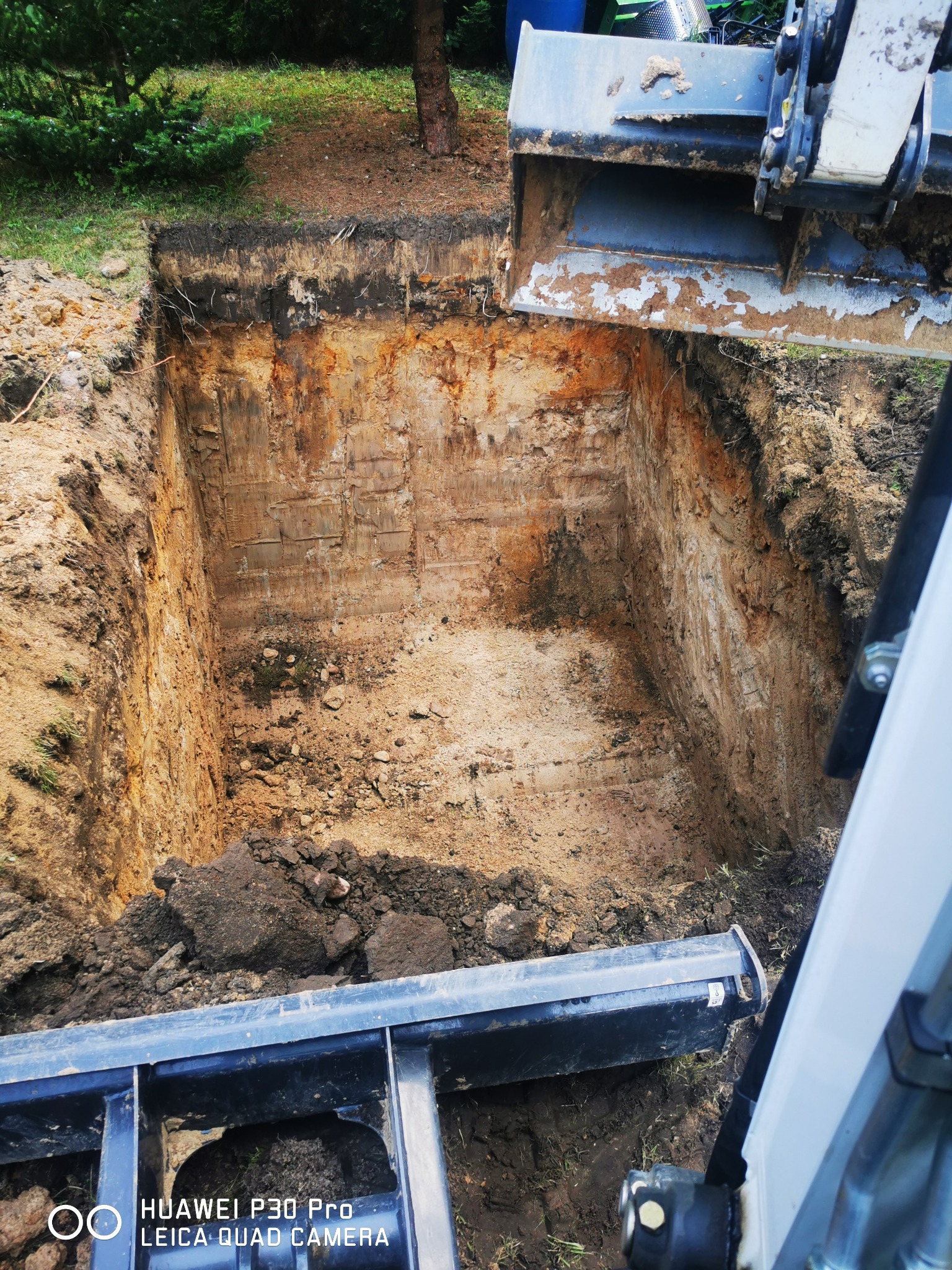 Głęboki wykop w ziemi, widoczna warstwowa struktura gleby, z boku łyżka koparki z resztkami ziemi, zielone otoczenie z drzewami w tle.