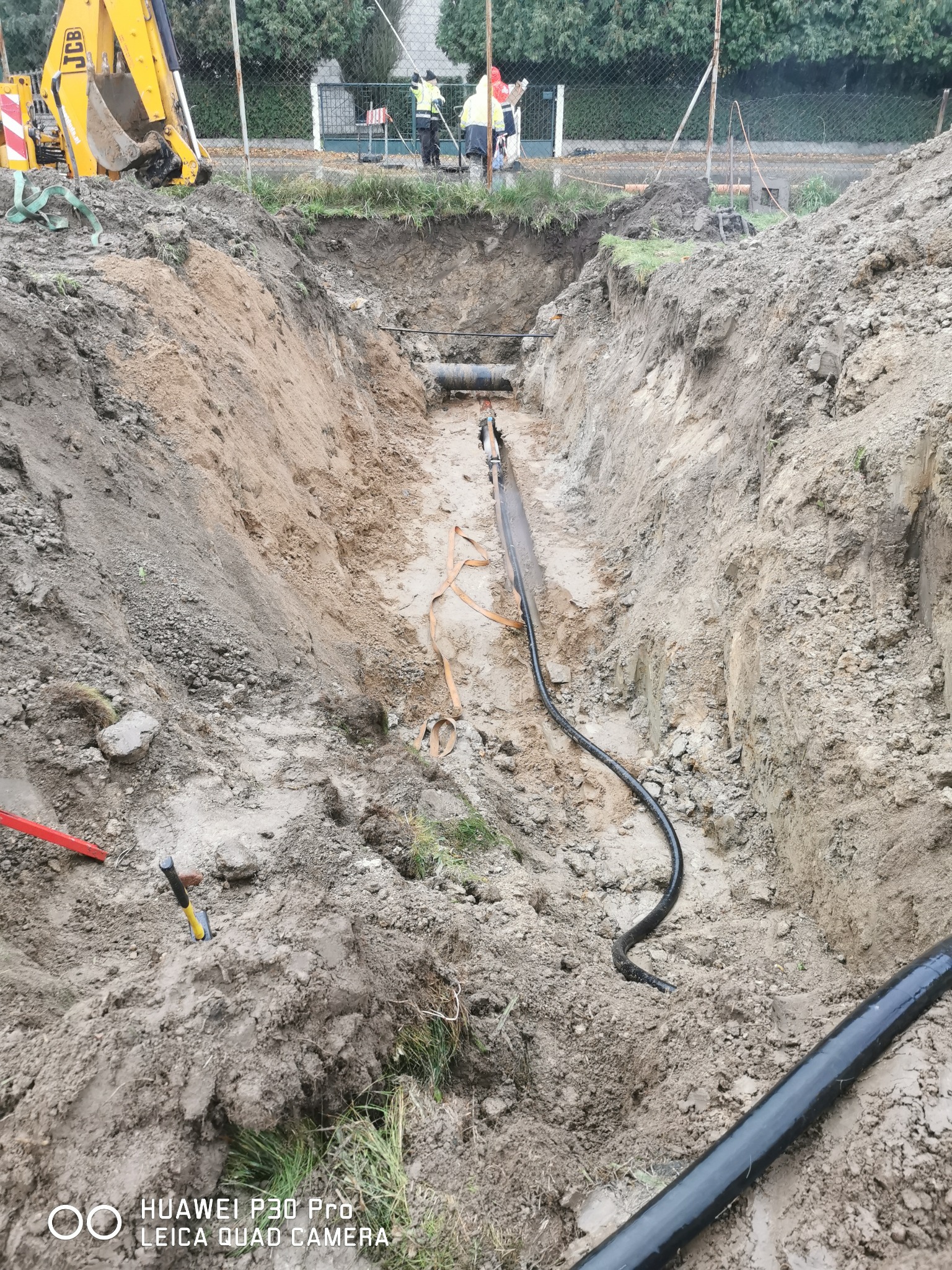 Głęboki wykop w ziemi z widocznymi rurami instalacyjnymi, żółta koparka JCB w tle, oraz pracownicy w odblaskowych kamizelkach w oddali.