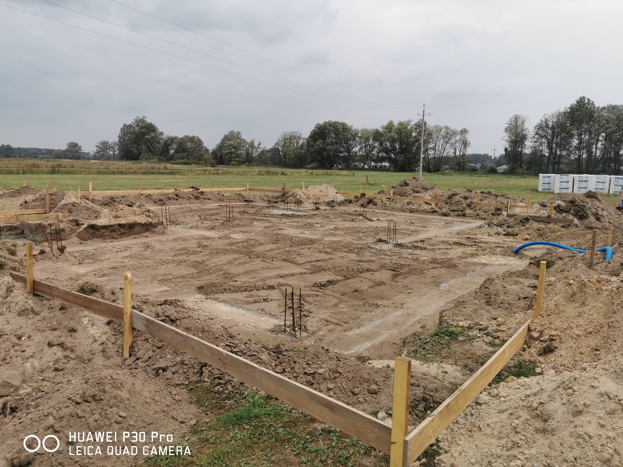 Przygotowany wykop pod fundamenty budynku z drewnianym szalunkiem, wystającymi prętami zbrojeniowymi i stertami bloczków Solbet w tle.