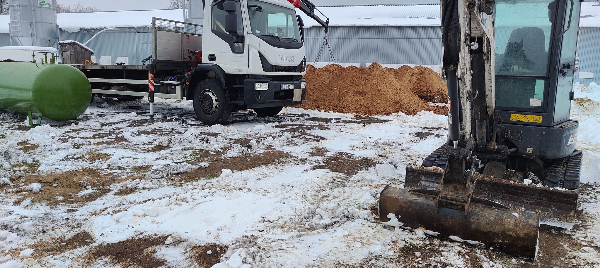 Mini koparka z łyżką na pierwszym planie, obok sterta ziemi, w tle biała ciężarówka Iveco z dźwigiem i zielony zbiornik, ziemia pokryta śniegiem.