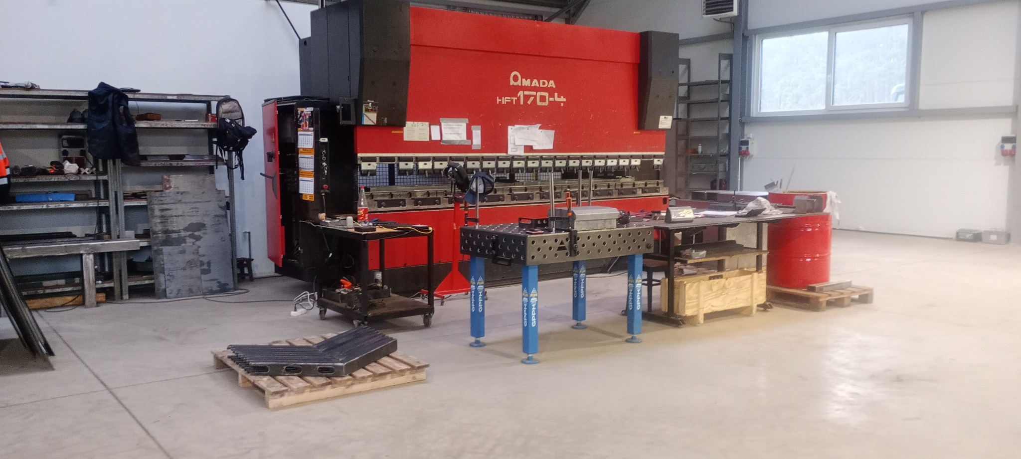 Czerwona prasa krawędziowa AMADA HFT 170-4 w hali produkcyjnej, obok stoliki warsztatowe, na podłodze paleta z elementami giętymi.