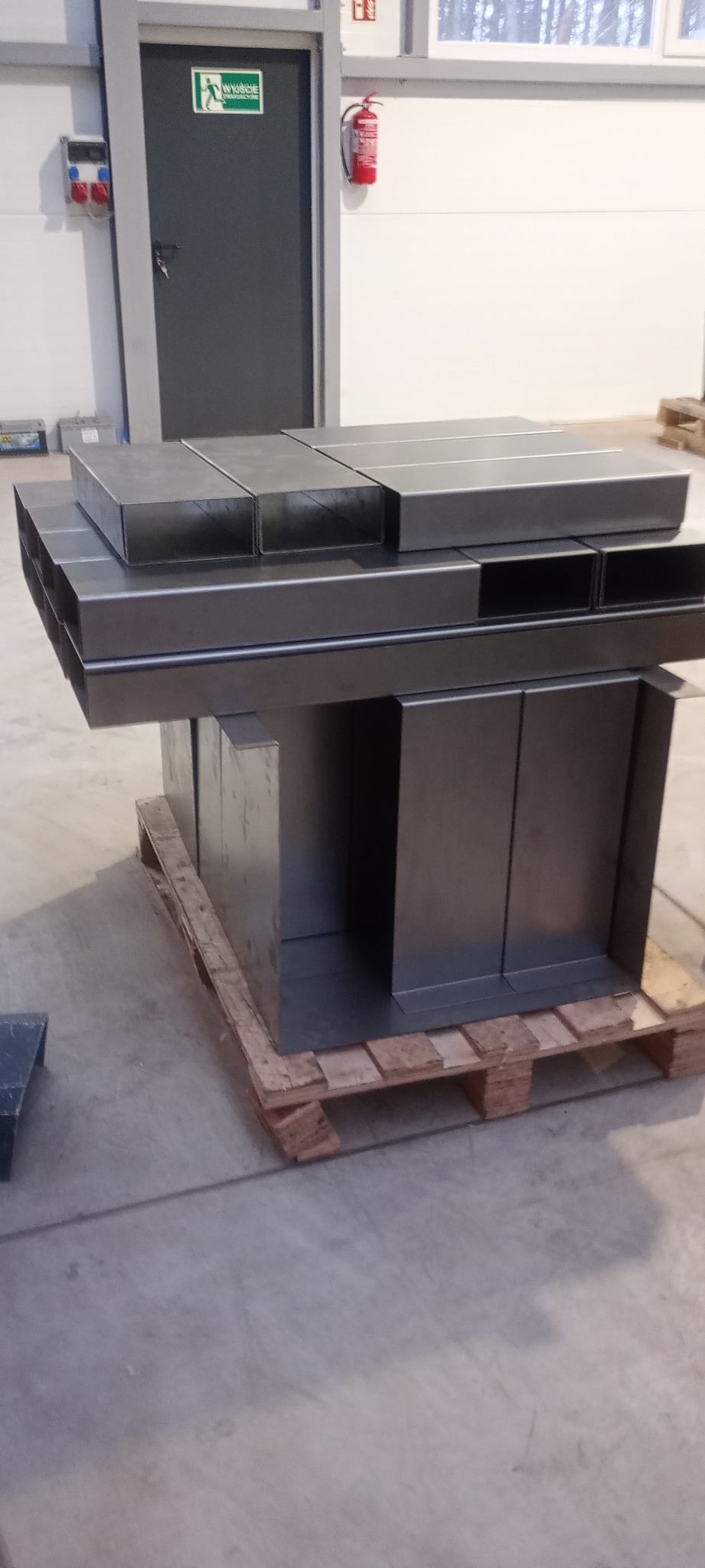 Metalowe prostokątne kanały wentylacyjne ułożone na palecie w magazynie, widoczne drzwi ewakuacyjne i gaśnica w tle.