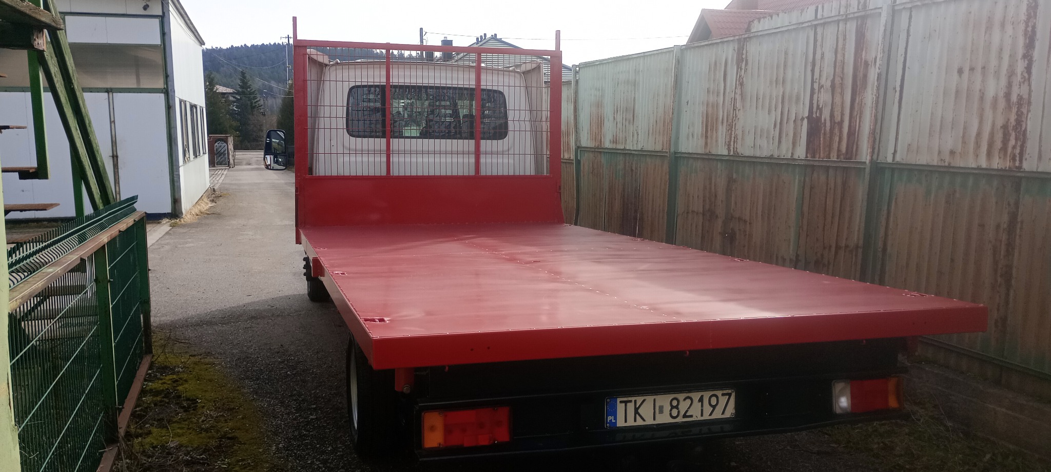 Czerwona platforma transportowa na podwoziu samochodu dostawczego, widok z tyłu, tablica rejestracyjna TKI 82197, tło: metalowe ogrodzenie i budynek.