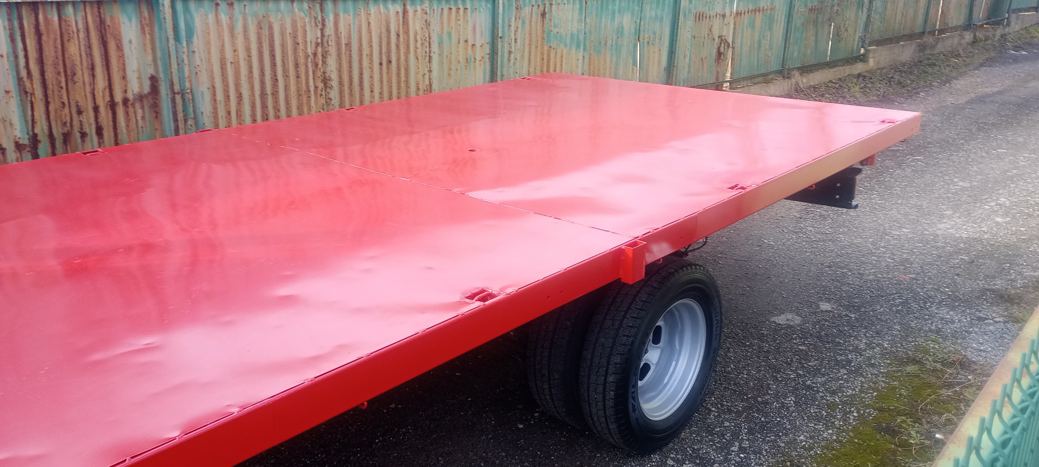 Czerwona, spawana platforma przyczepy samochodowej z podwójnymi kołami, stojąca na zewnątrz na tle zardzewiałego ogrodzenia.