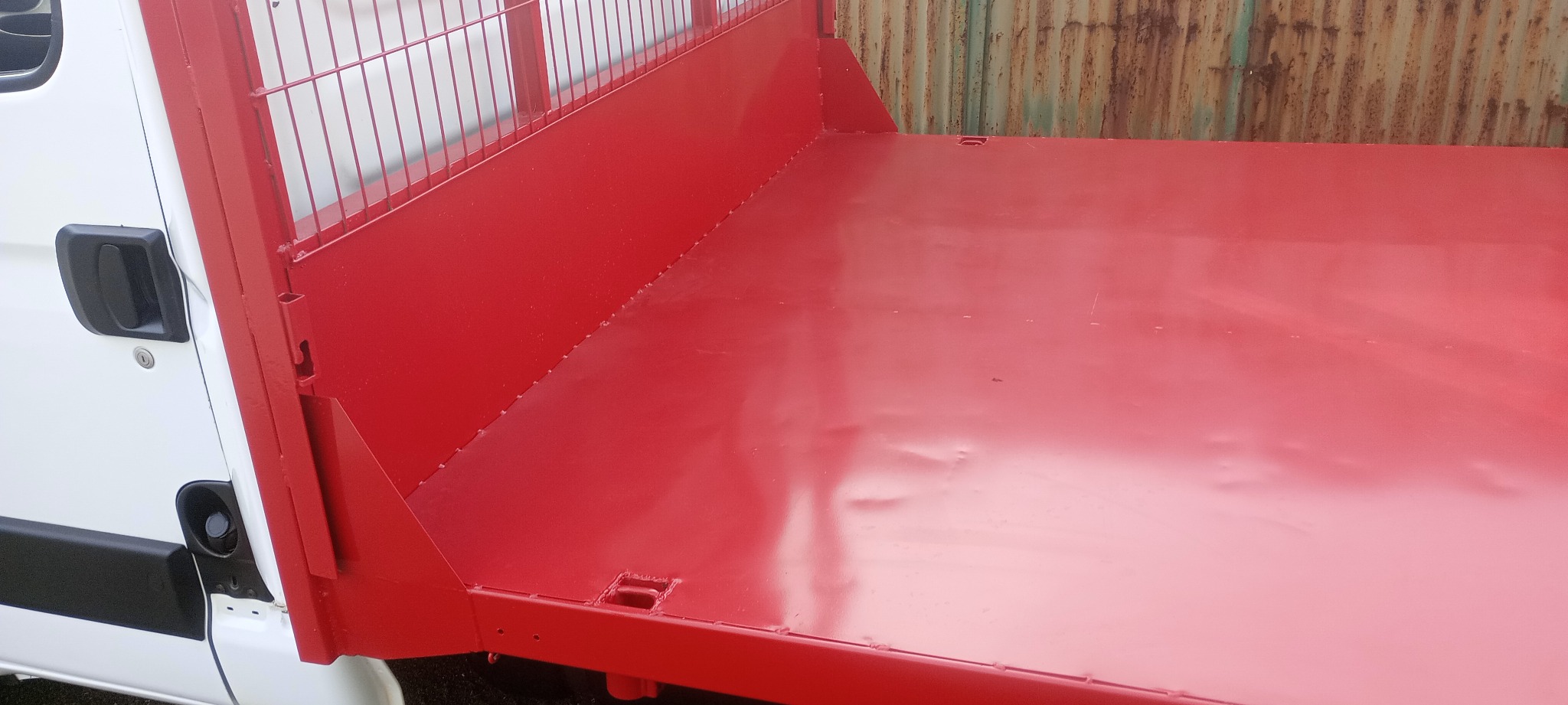 Czerwona, metalowa skrzynia ładunkowa samochodu dostawczego, widok z boku, z widocznymi elementami konstrukcyjnymi i fragmentem białej kabiny.