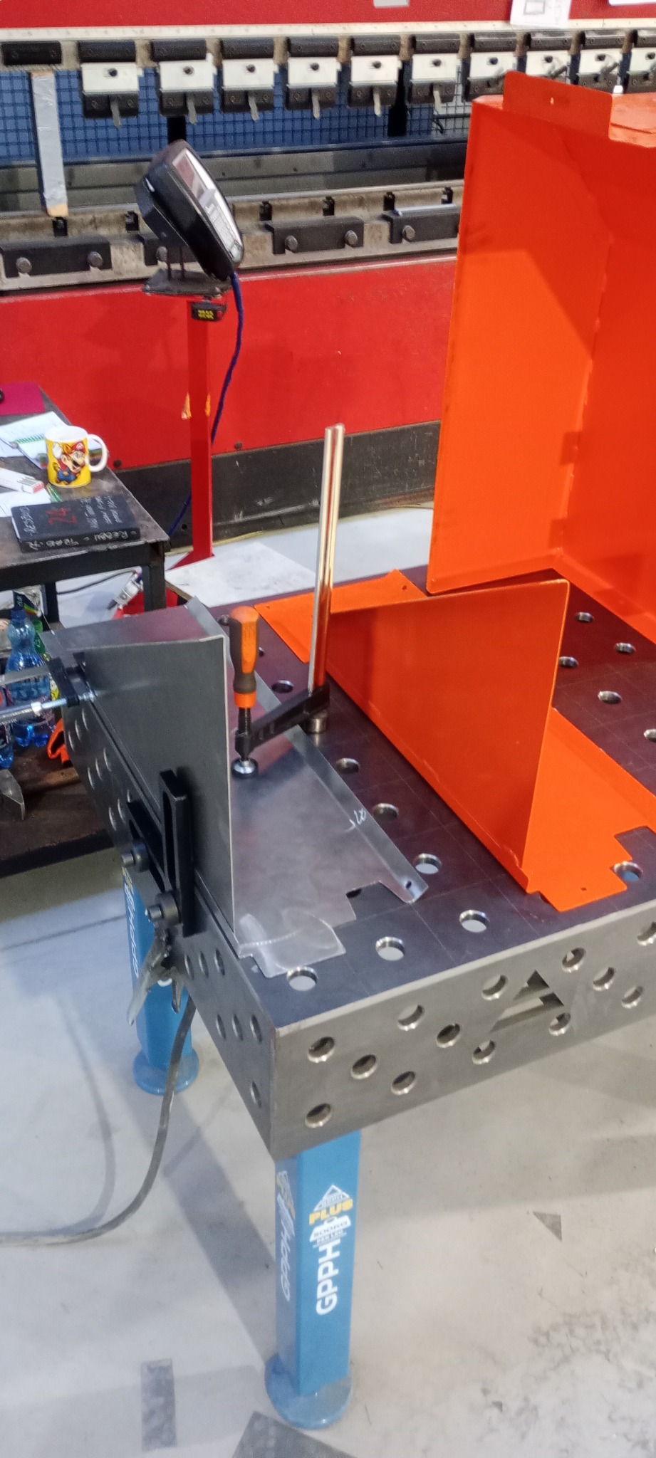 Metalowe elementy, zespawane pod kątem prostym, zamocowane na perforowanym stole spawalniczym z użyciem ścisków i kątownika, w tle czerwona maszyna do obróbki blach.