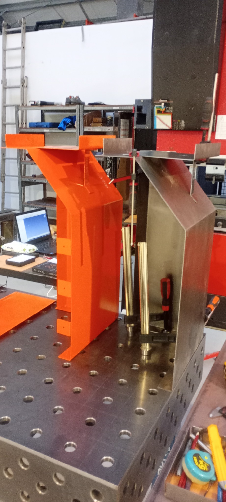 Dwa metalowe elementy (pomarańczowy i srebrny) zamocowane na perforowanym stole spawalniczym, widoczne narzędzia i laptop w tle.