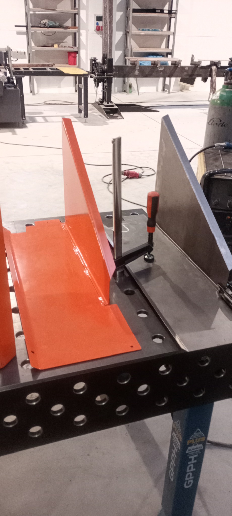 Pomarańczowy i szary element metalowy z widocznym spoiwem na perforowanym stole warsztatowym, w tle sprzęt spawalniczy i regały.