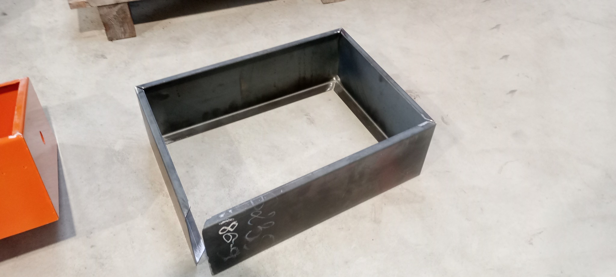 Prostokątna, metalowa konstrukcja spawana z blachy, z widocznymi śladami po spawaniu i oznaczeniami na boku, ustawiona na betonowej posadzce, obok pomarańczowego elementu.