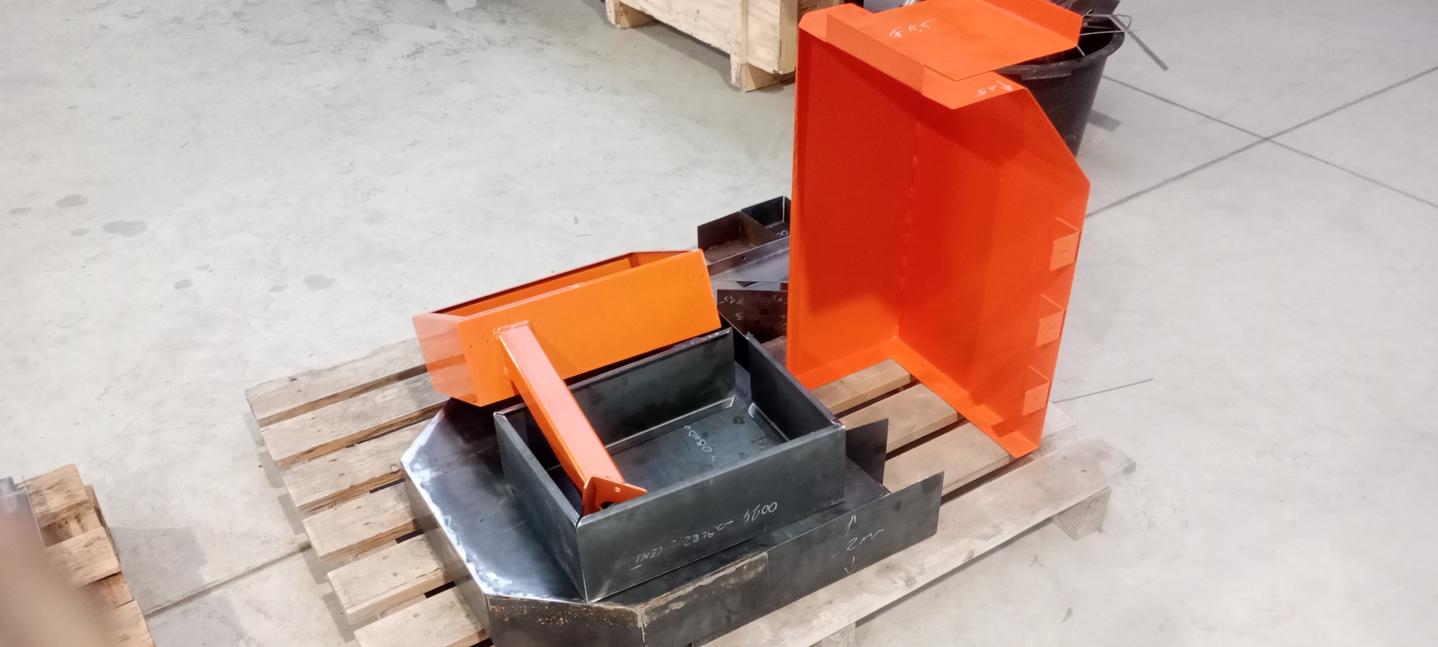 Pomarańczowe i czarne metalowe elementy spawane, niektóre pomalowane, ułożone na palecie w hali produkcyjnej.