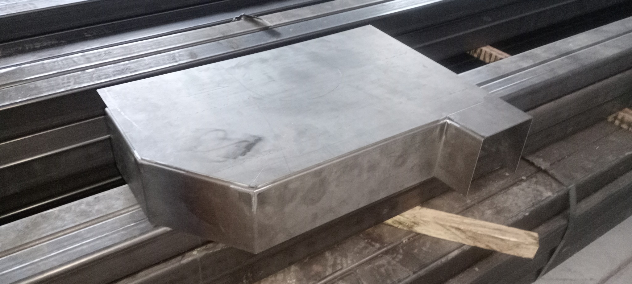 Metalowa konstrukcja o nieregularnym kształcie, z widocznymi spawami, leżąca na stosie profili stalowych.