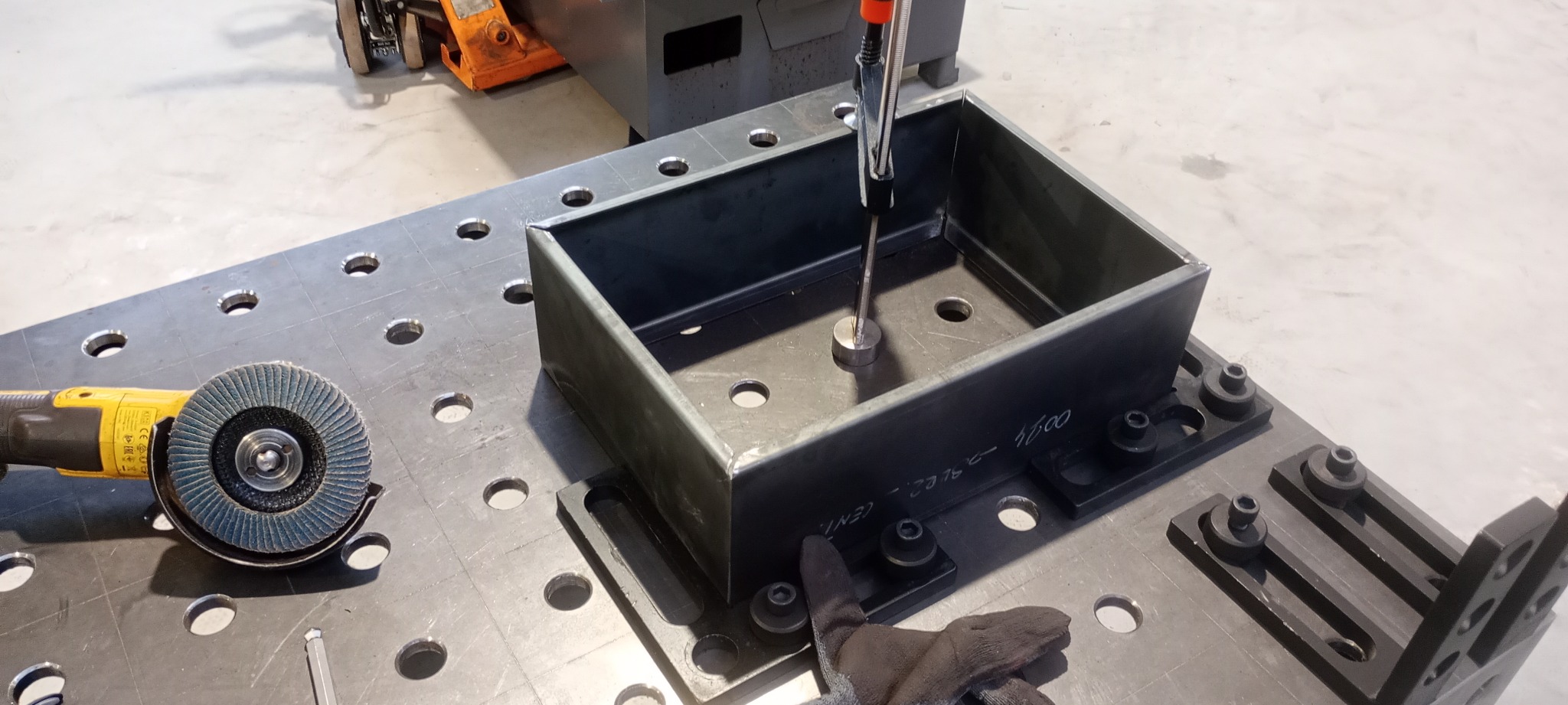 Metalowa konstrukcja skrzynkowa, częściowo zespawana, umocowana na stole spawalniczym z otworami, obok szlifierka kątowa z tarczą listkową i rękawice robocze.