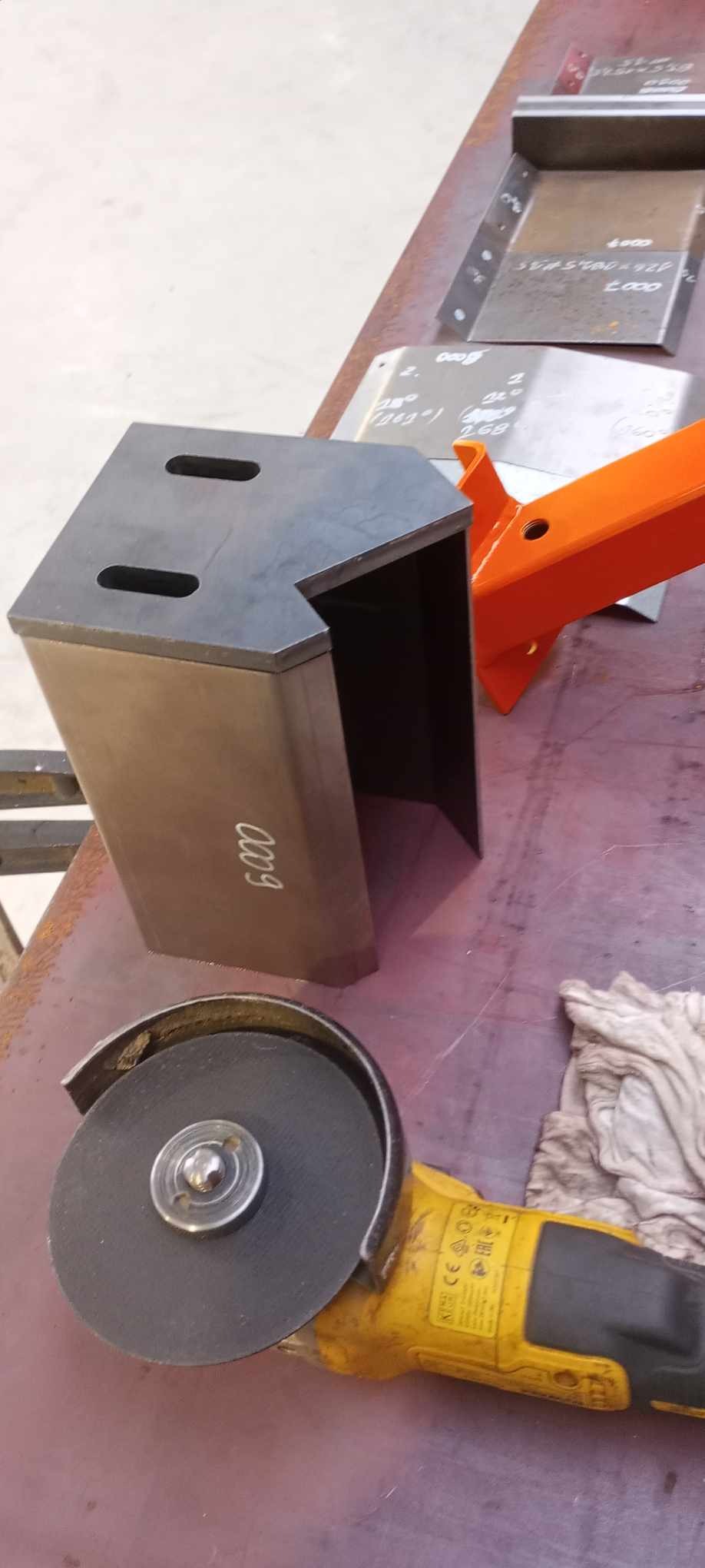 Metalowa konstrukcja z wyciętymi otworami, obok szlifierka kątowa i pomalowany na pomarańczowo element metalowy, na stole warsztatowym.