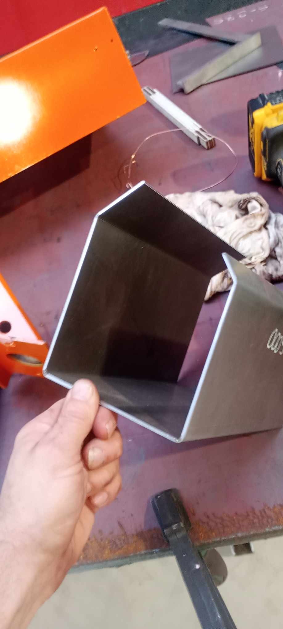 Metalowy profil kątowy trzymany w dłoni, widoczne narzędzia i elementy warsztatu w tle, w tym pomarańczowy element konstrukcyjny.