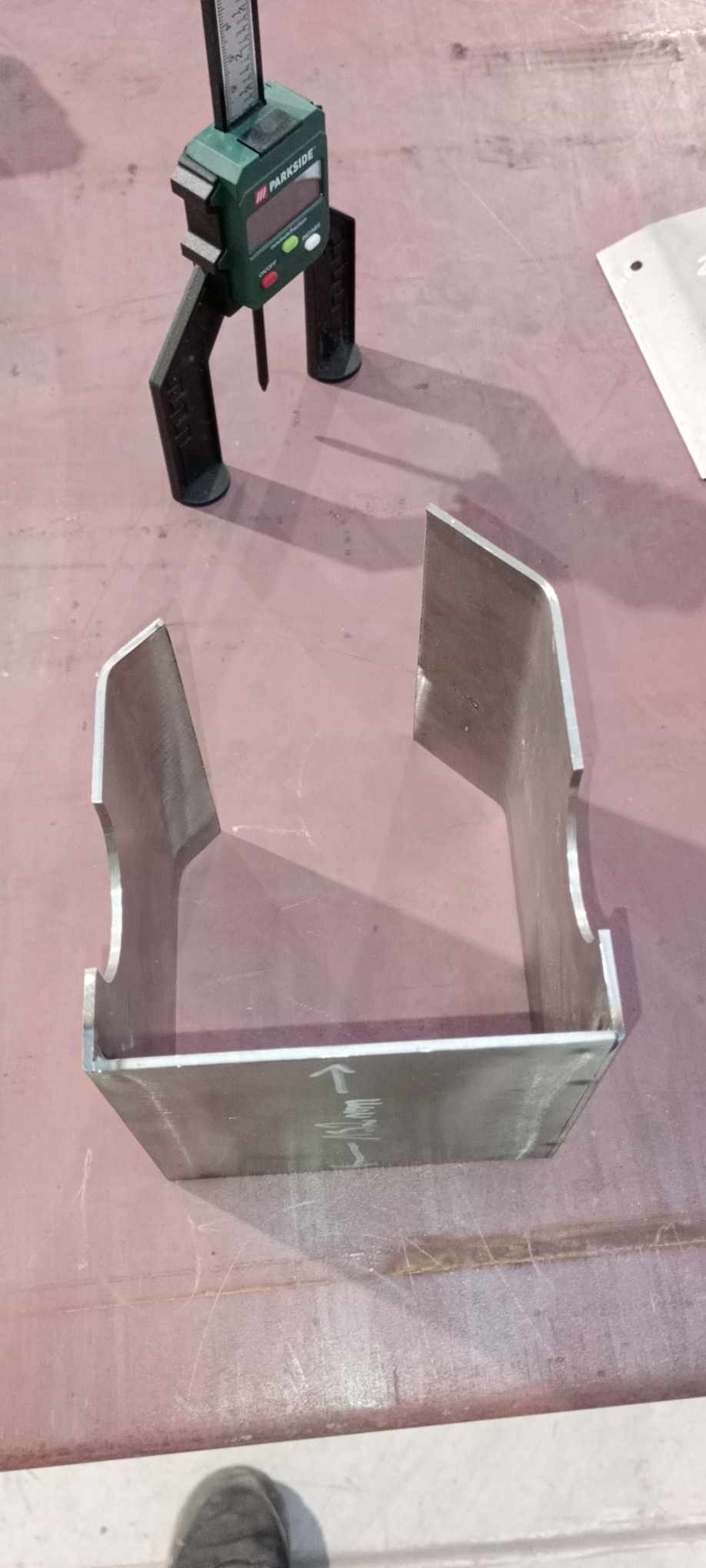 Metalowy element z wycięciami, sprawdzany suwmiarką elektroniczną Parkside, leżący na brudnym metalowym stole.