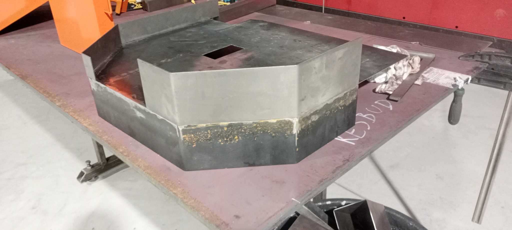 Metalowa konstrukcja spawana o nietypowym kształcie, z widocznymi śladami spawania i obróbki, umieszczona na stole warsztatowym z narzędziami i zabrudzeniami.