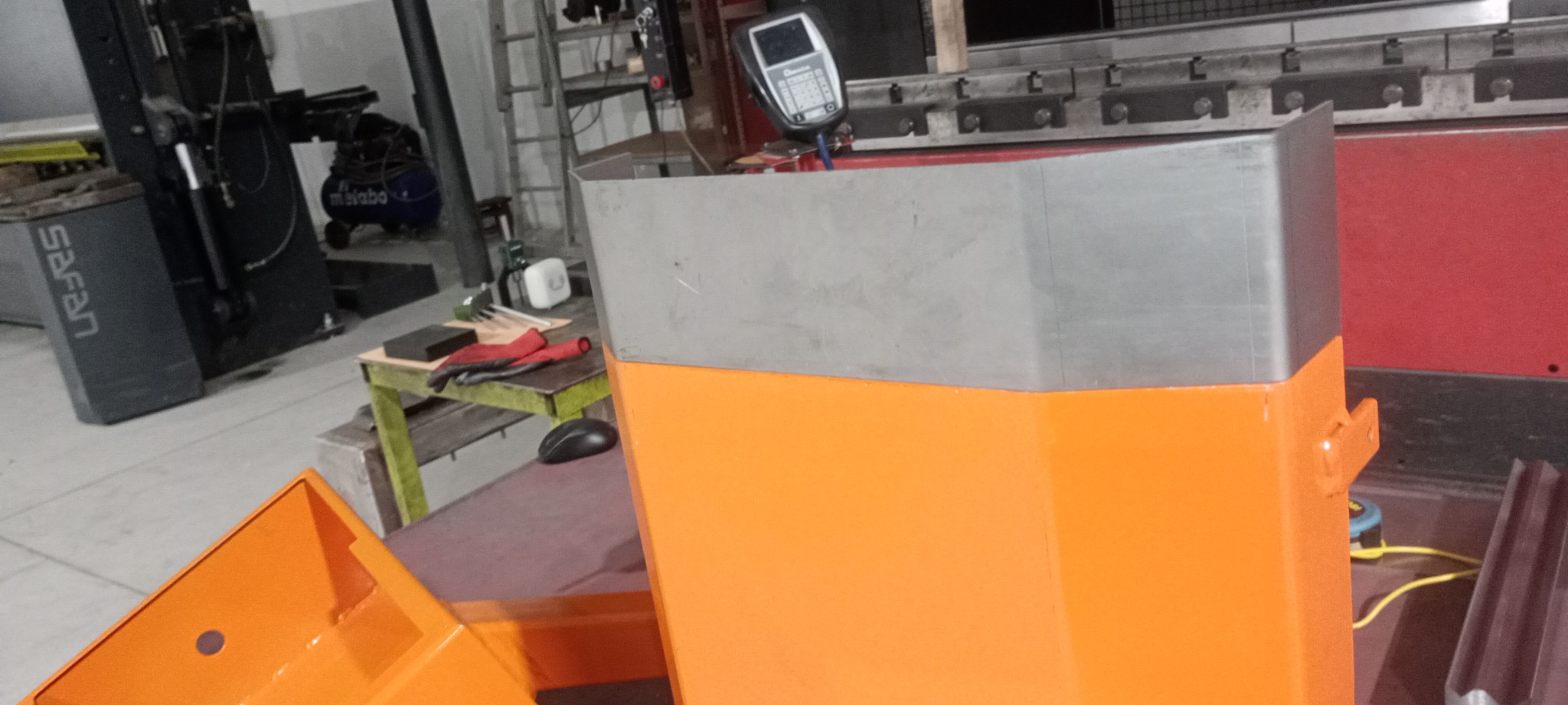 Pomarańczowo-szara, metalowa konstrukcja po obróbce, z widocznymi śladami spawania i obróbki blacharskiej, na tle warsztatu z narzędziami i maszynami.