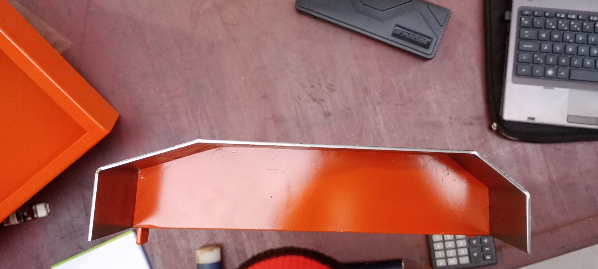 Pomarańczowa metalowa konstrukcja z widocznymi śladami spawania, umieszczona na stole obok laptopa i kalkulatora.
