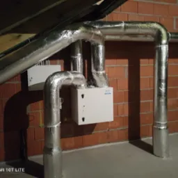 Centrala rekuperacyjna zamontowana na poddaszu, z widocznymi izolowanymi przewodami wentylacyjnymi i ceglaną ścianą w tle.