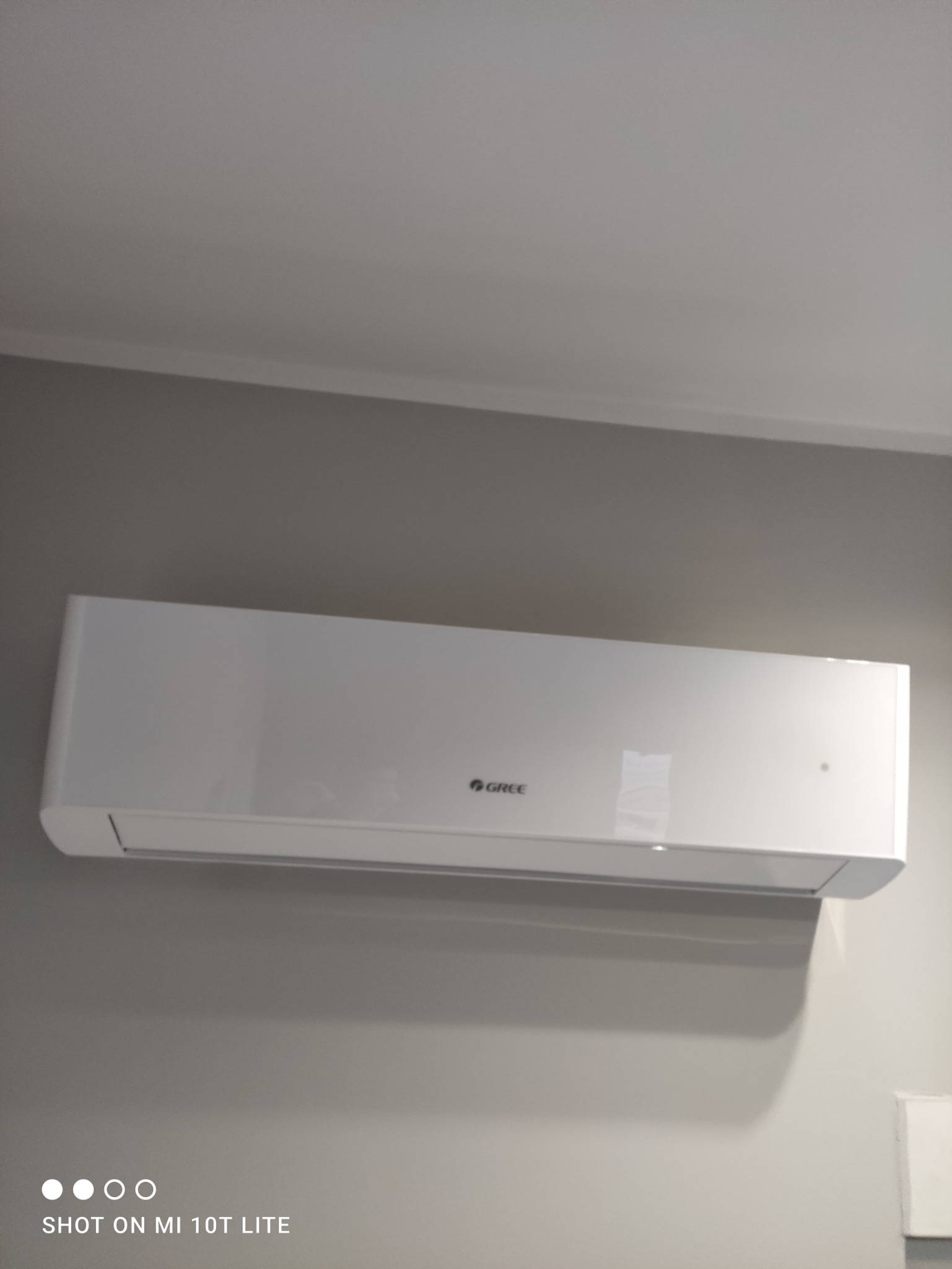 Biała jednostka klimatyzacji marki Gree zamontowana na szarej ścianie, widok z dołu.