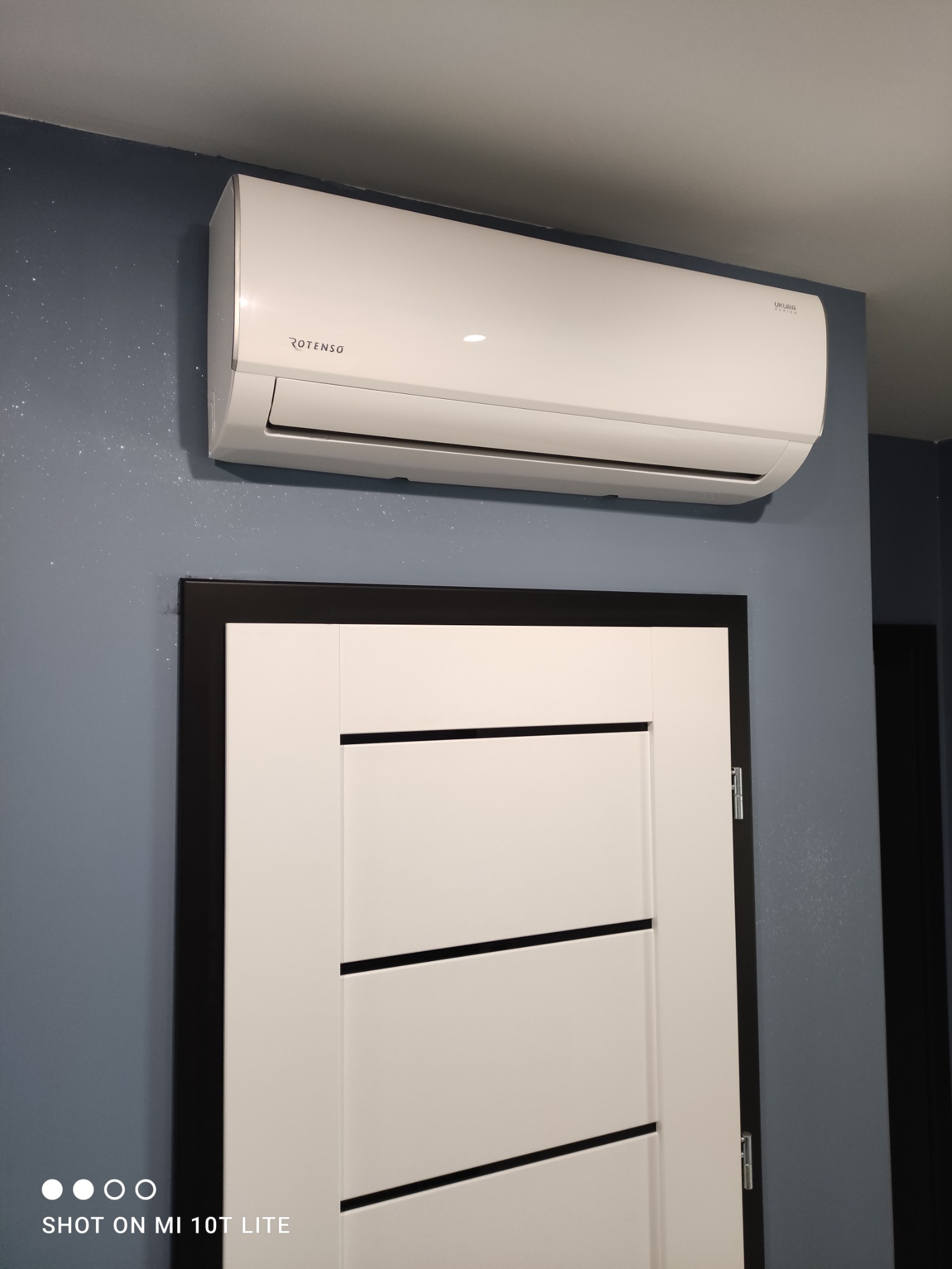 Biały klimatyzator marki Rotenso zamontowany nad nowoczesnymi, białymi drzwiami z czarnymi akcentami, na tle ściany w kolorze gołębiej szarości z subtelnym połyskiem.