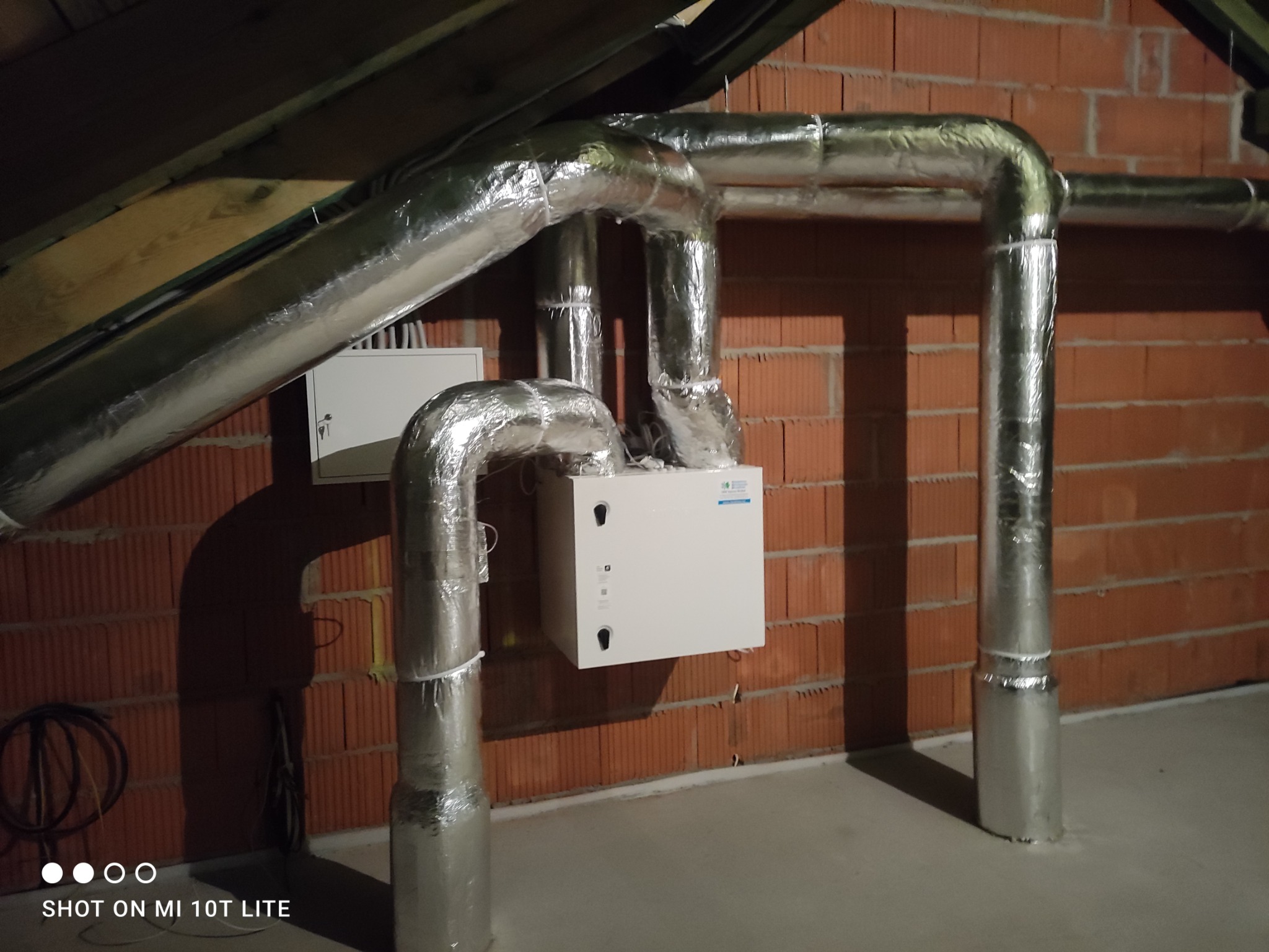 Centrala rekuperacyjna zamontowana na poddaszu, z widocznymi izolowanymi przewodami wentylacyjnymi i ceglaną ścianą w tle.