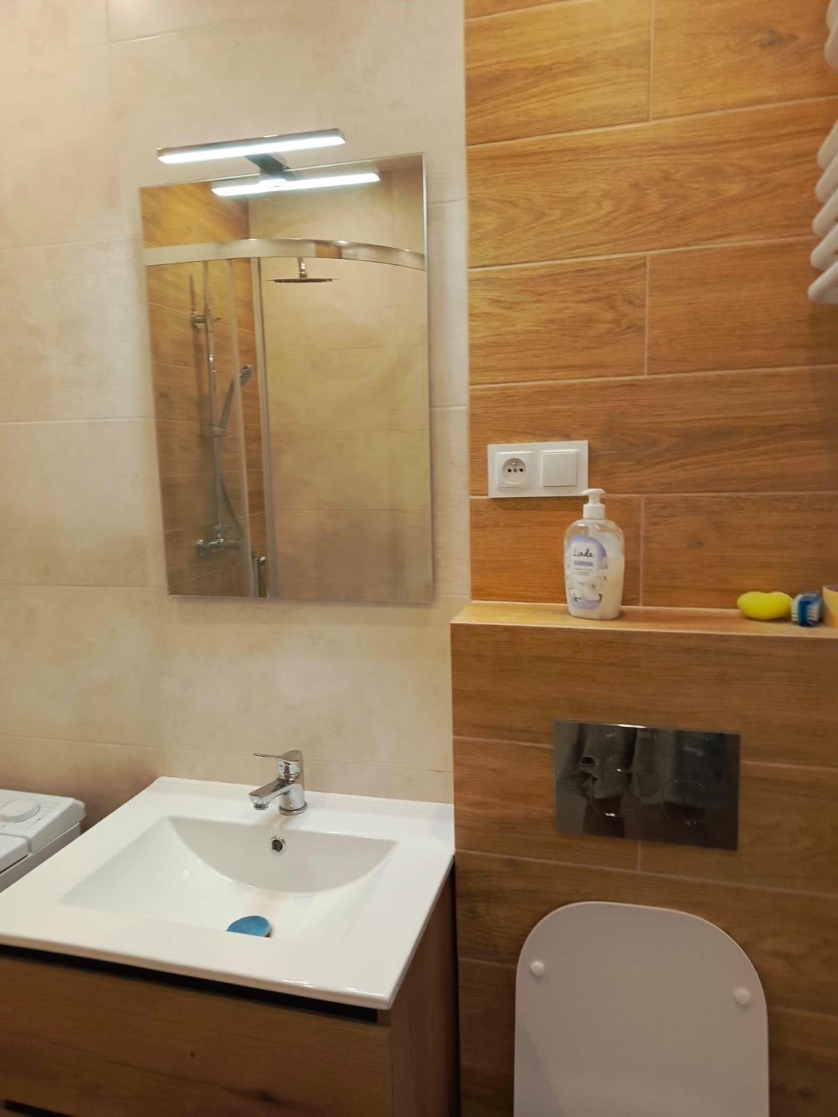 Jasna łazienka z drewnopodobnymi płytkami, umywalką z szafką i lustrem z oświetleniem. W tle widoczna kabina prysznicowa. Minimalistyczny design.