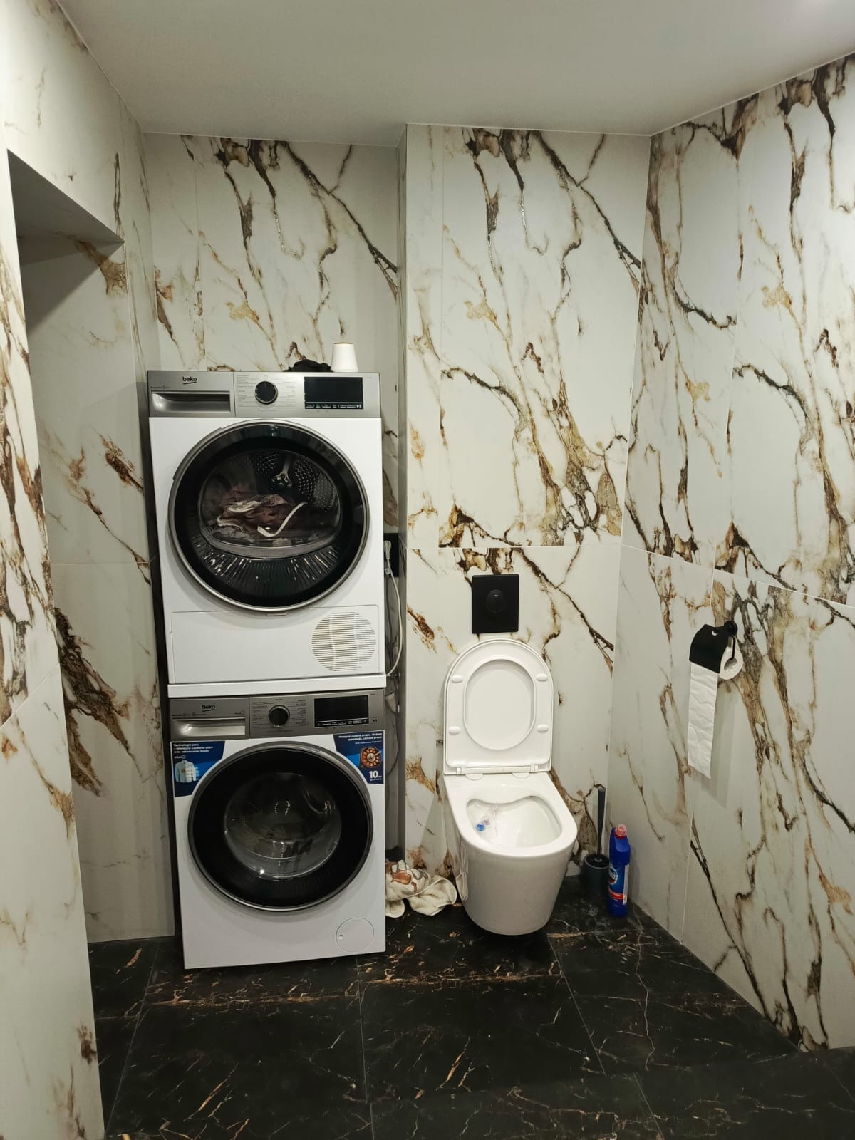 Wykończona łazienka z marmurowymi płytkami, toaletą i pralko-suszarką Beko ustawioną w pionie. Ciemna podłoga z żyłkowaniem. Widok na wnętrze łazienki.