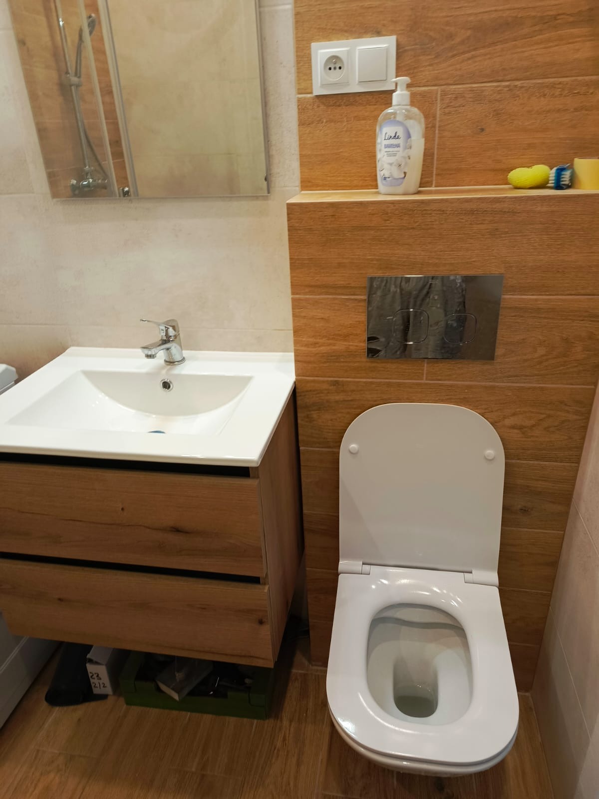 Nowoczesna łazienka z drewnopodobnymi płytkami, umywalką z szafką i podwieszaną toaletą. Widoczny fragment lustra i akcesoria łazienkowe.