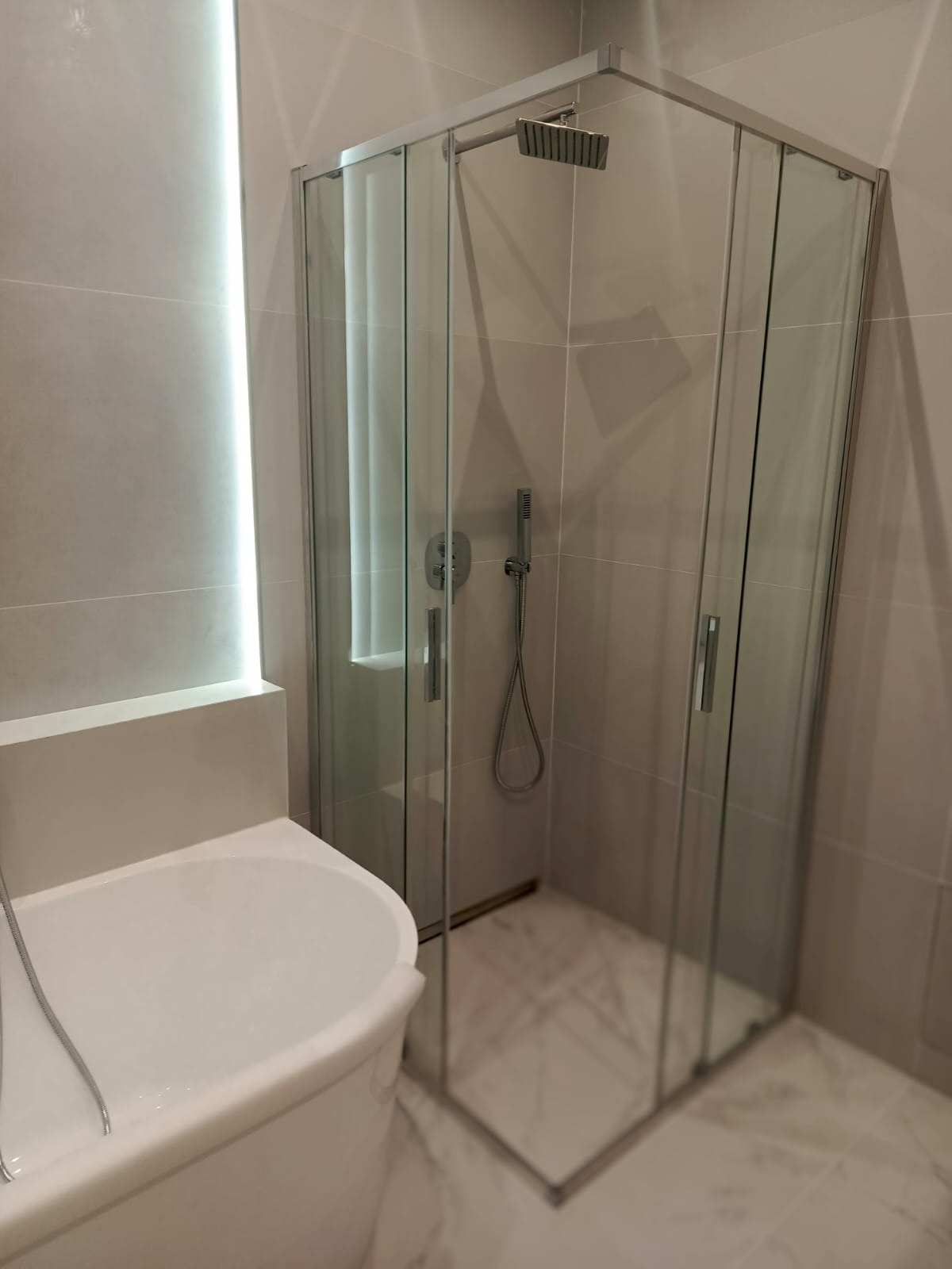 Nowoczesna łazienka z kabiną prysznicową typu walk-in i wolnostojącą wanną. Minimalistyczny design, jasne płytki, subtelne oświetlenie LED.