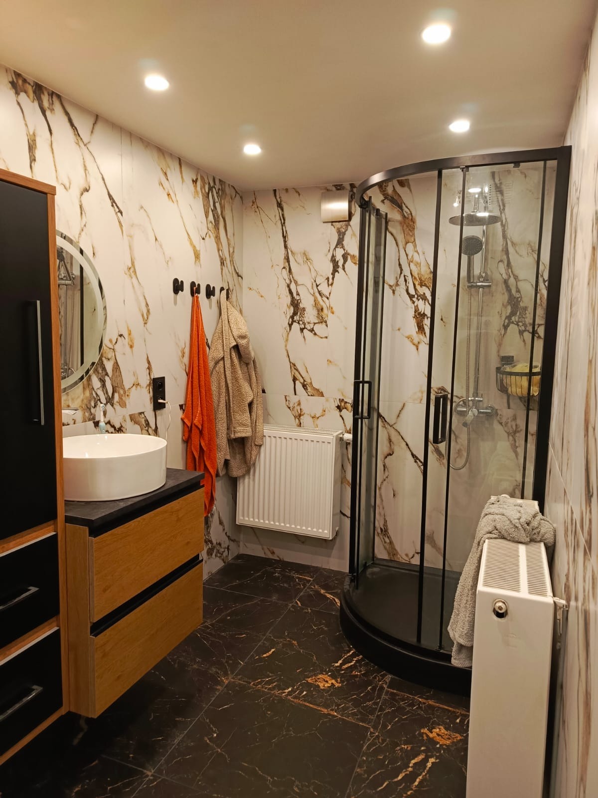 Nowoczesna łazienka z kabiną prysznicową typu walk-in, umywalką nablatową i marmurowymi płytkami. Widoczny grzejnik i akcesoria łazienkowe.