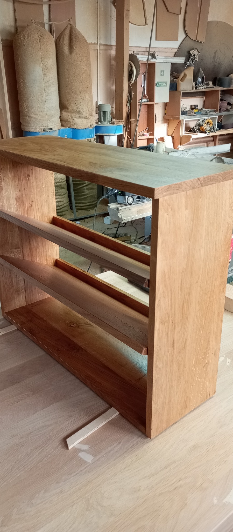 Surowa drewniana szafka z półkami w warsztacie stolarskim, widoczne worki na trociny i narzędzia w tle.
