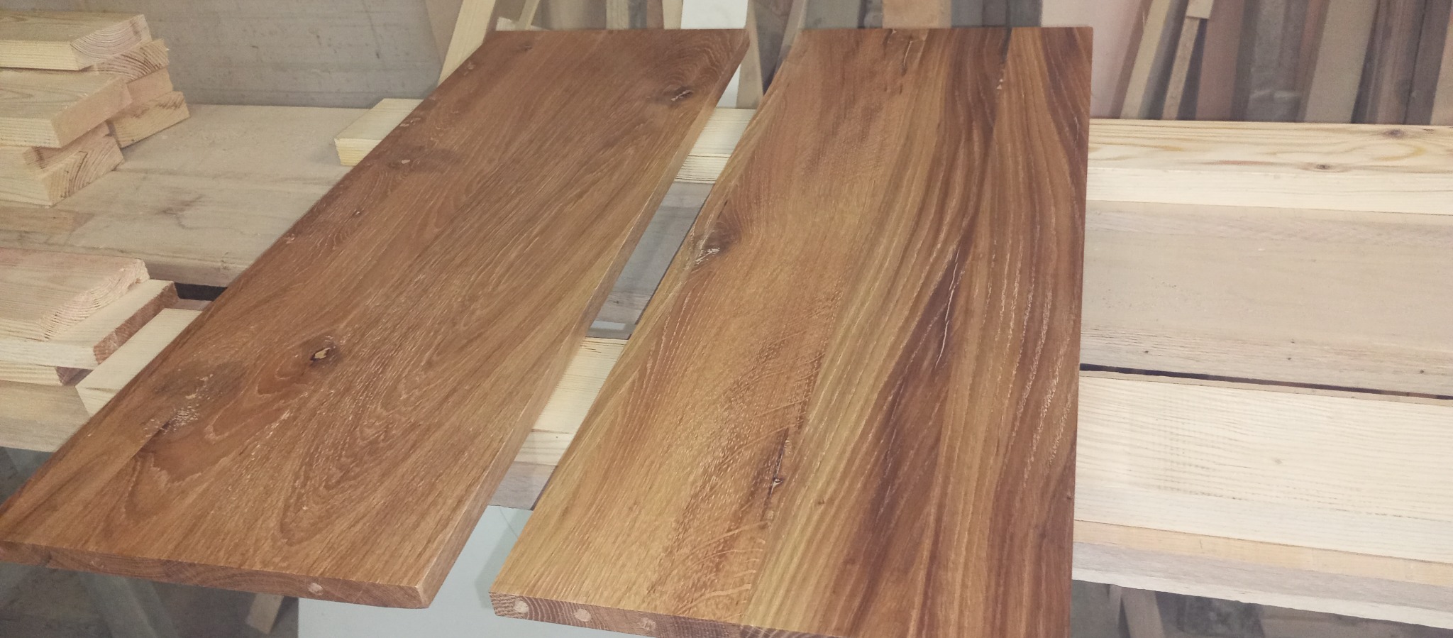 Dwie surowe deski z drewna liściastego o wyraźnym usłojeniu, leżące obok siebie na drewnianym regale warsztatowym.