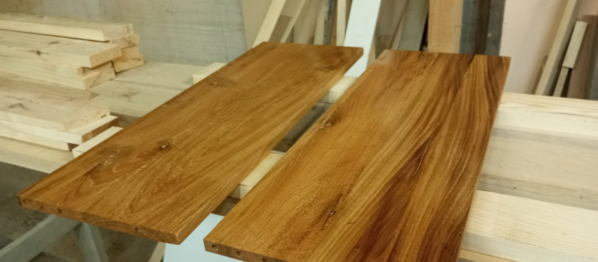 Dwie deski z jasnego drewna o wyraźnym usłojeniu, leżące na stosie innych drewnianych elementów w warsztacie stolarskim.